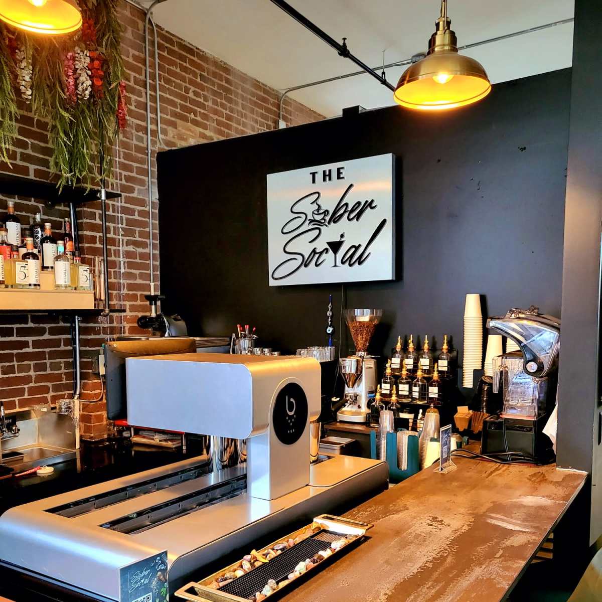 A modern coffee bar with a sleek espresso machine, a variety of bottles and equipment on display, and a dark brick wall adorned with decorative plants.