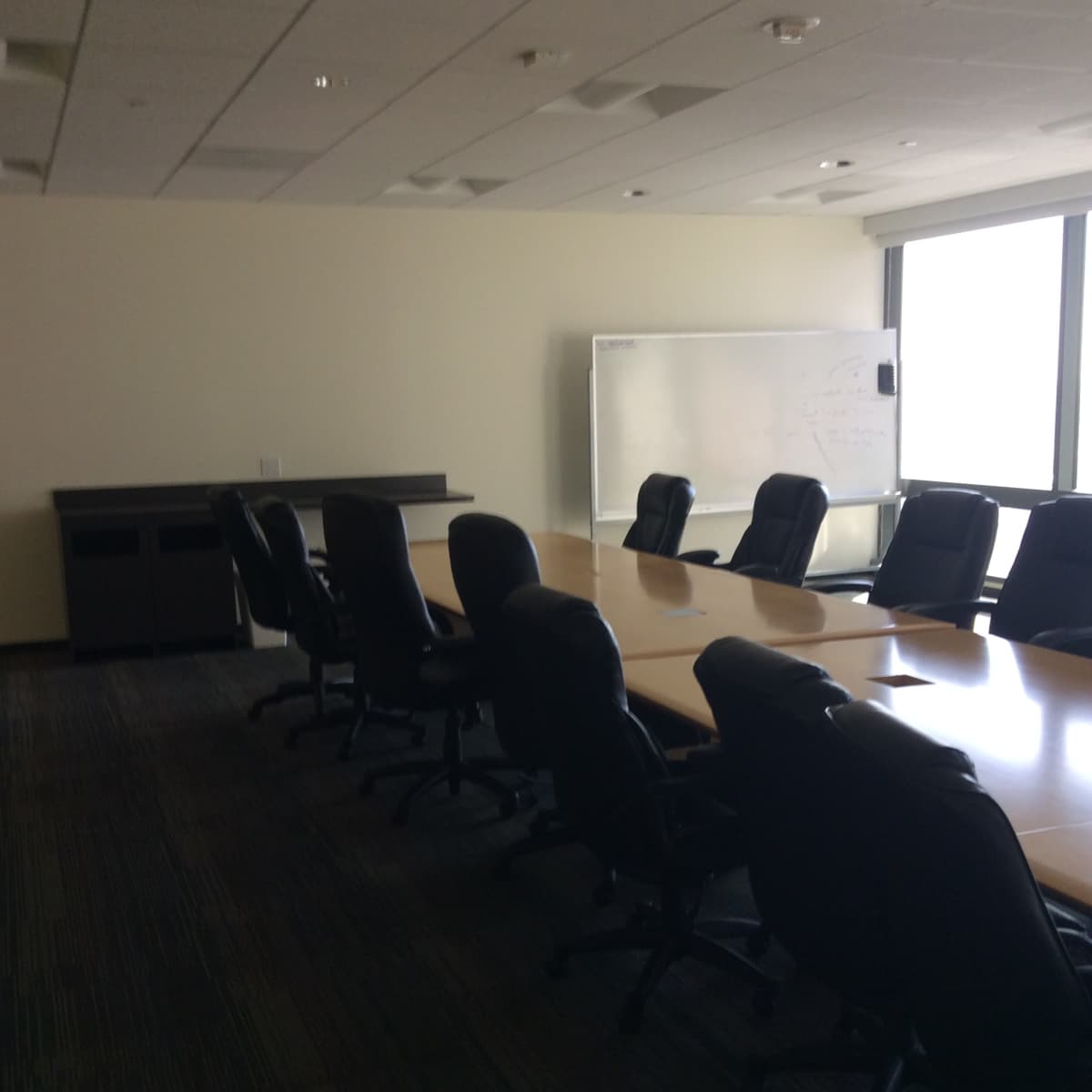 A conference room features a long table surrounded by black chairs, with a whiteboard positioned against a wall and large windows allowing natural light.