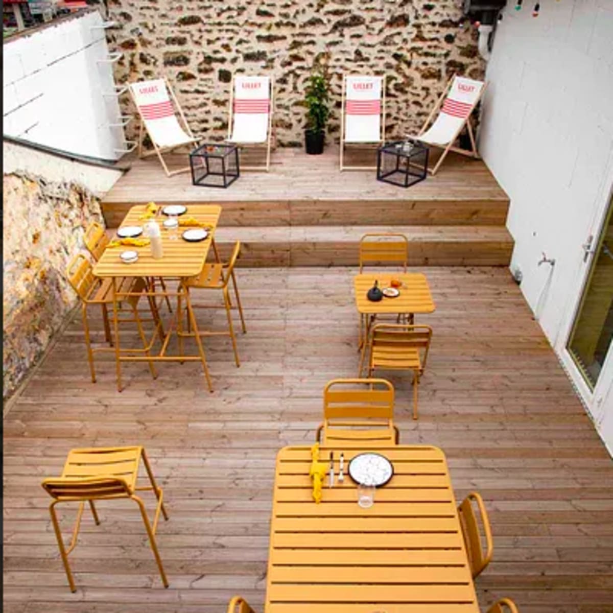 A wooden deck area with several yellow metal tables and chairs, accompanied by deck chairs in the background against a stone wall.