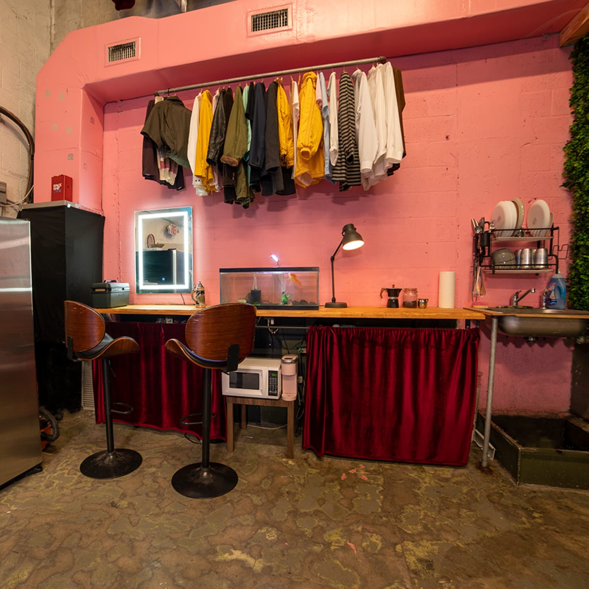 A small room featuring a pink wall with hanging clothes, a stainless steel refrigerator, a wooden countertop with bar stools, a mirror, and a small microwave.