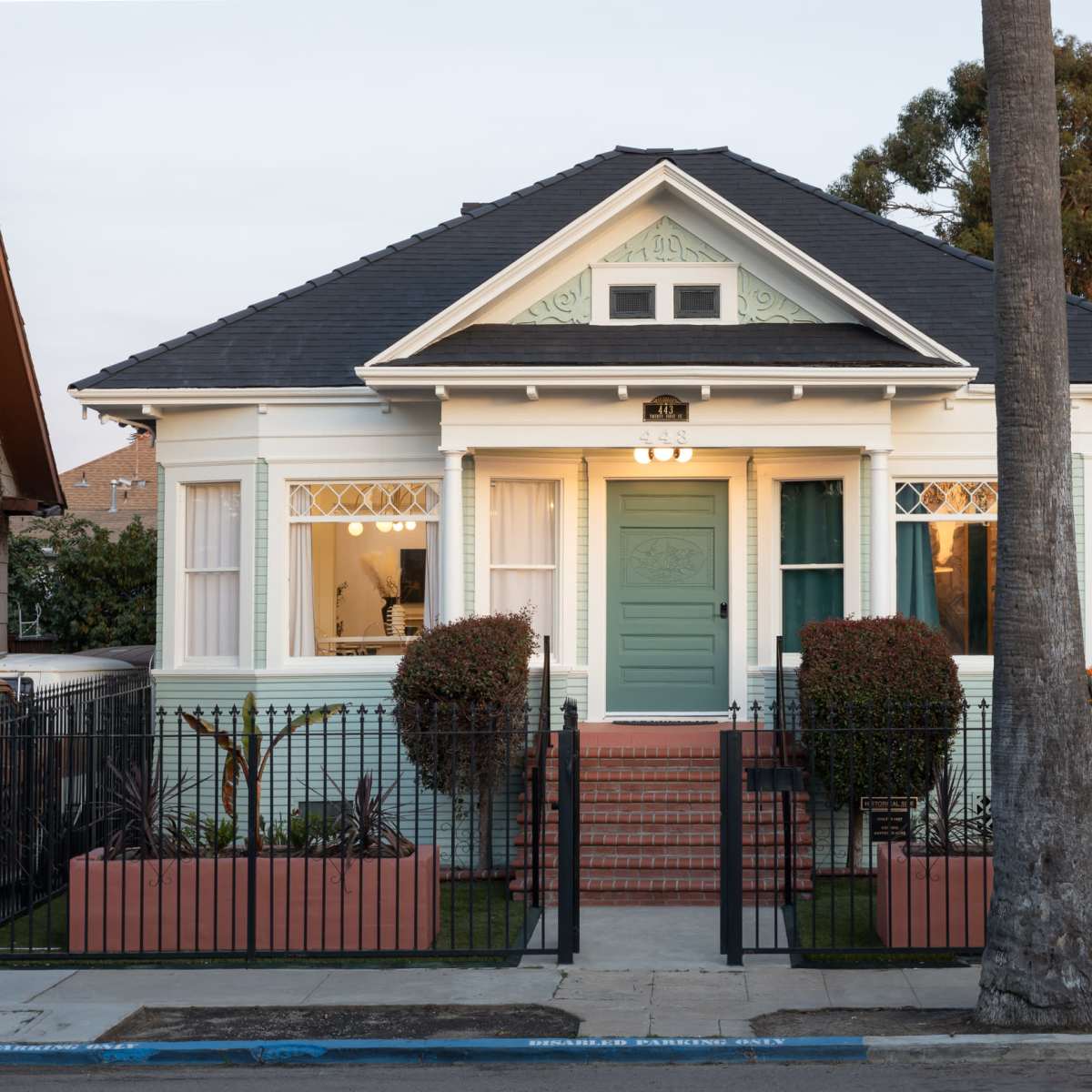 A single-story house with a light blue door, surrounded by a black fence and neatly arranged landscaping, including two bushy plants in front.
