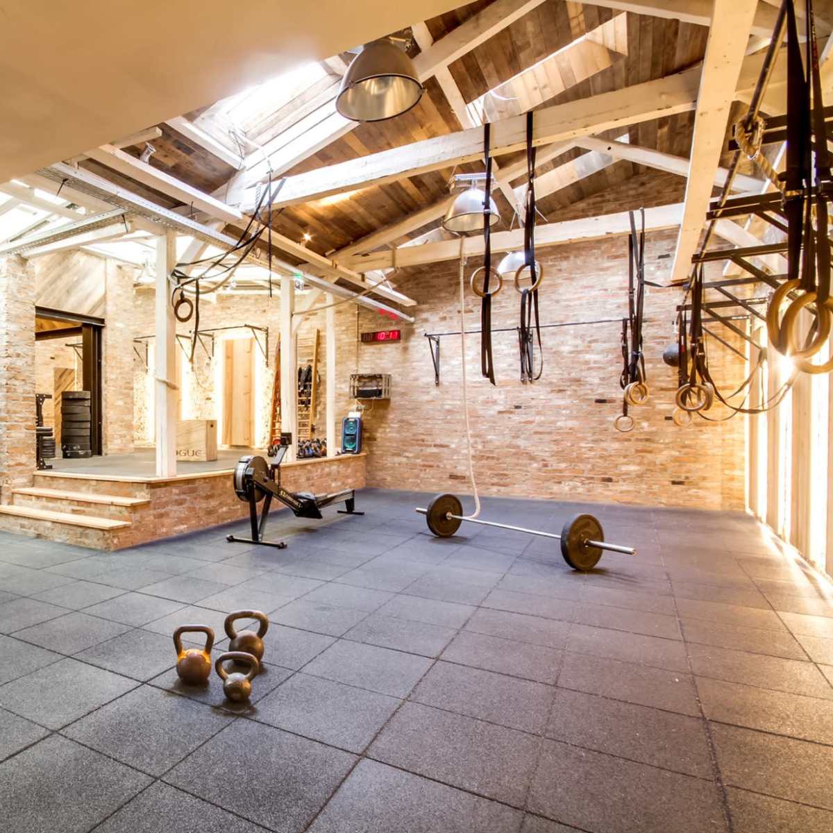 A spacious, industrial-style gym with exposed brick walls, various weightlifting equipment, and fitness tools arranged on a rubber floor.