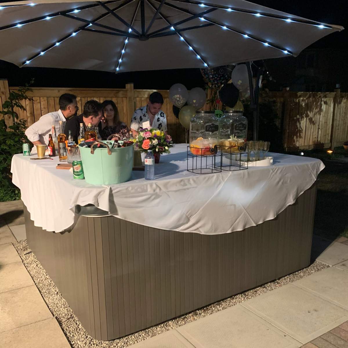 A group of people gather around a bar setup with drinks and decorations under an umbrella in a backyard setting at night.