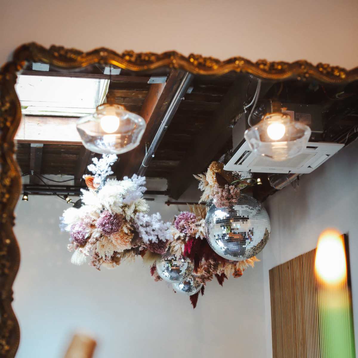 A decorative mirror reflects a ceiling adorned with floral arrangements and disco balls, with a candle in the foreground.