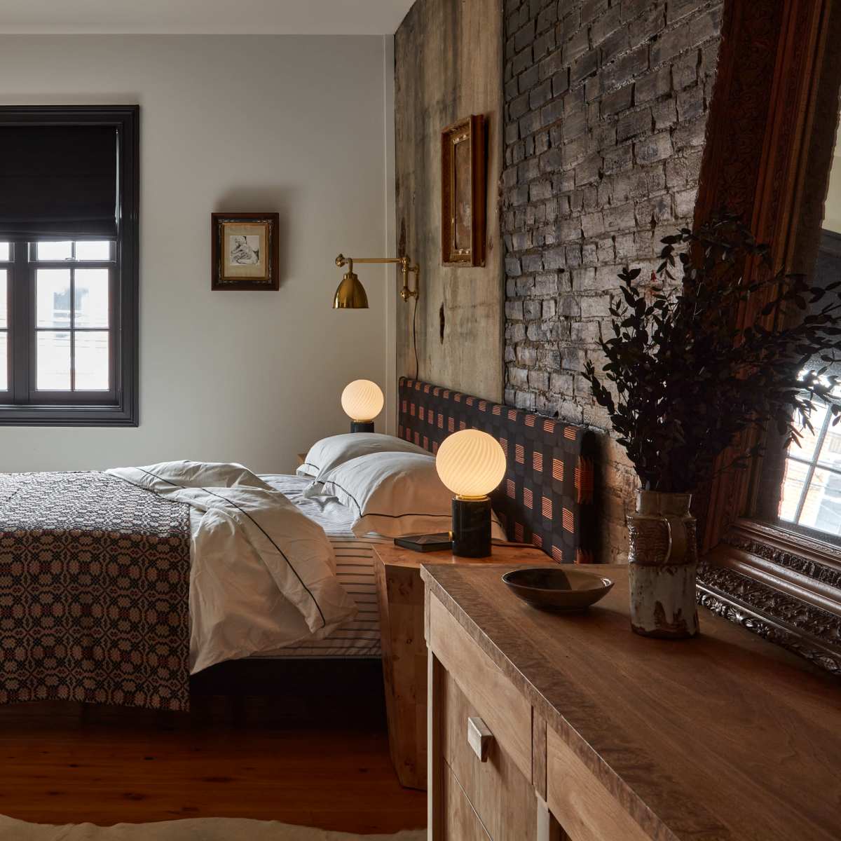 A bedroom featuring a bed with a patterned quilt, a wooden dresser, and large windows with blinds, set against a brick wall.