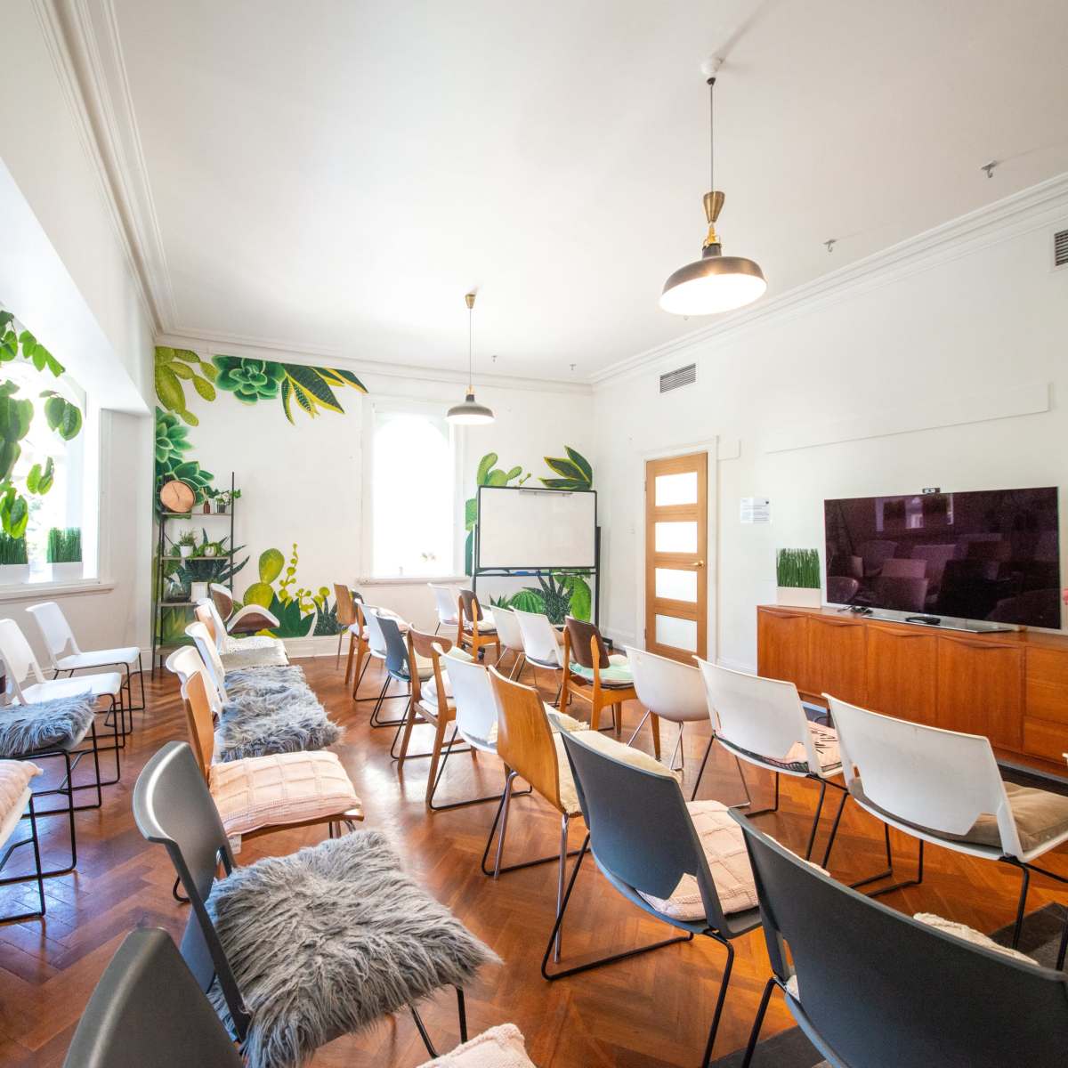 The room features arranged chairs and a large television, with plants painted on the walls and wooden accents throughout.