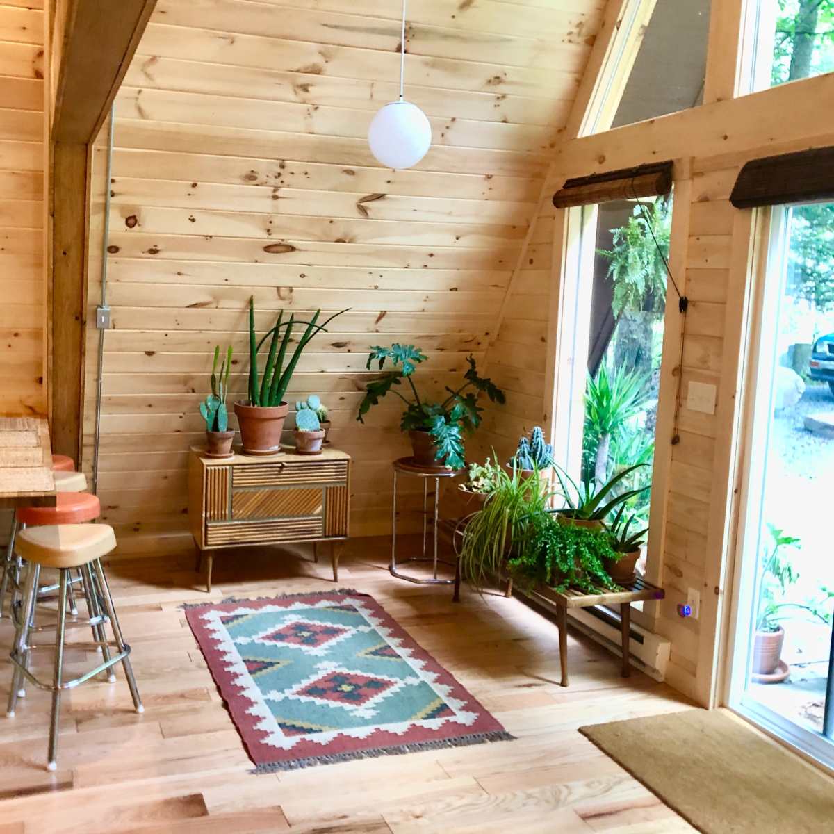 A wooden interior featuring an A-frame design, with a small dining area, a rug with geometric patterns, potted plants, and large windows providing natural light.