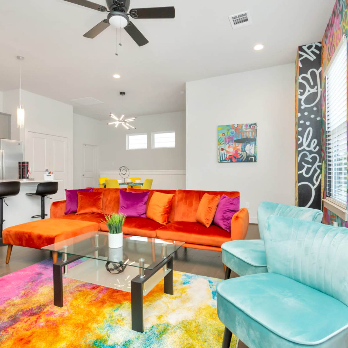 A brightly decorated living area with a colorful sofa, glass coffee table, and stylish chairs, adjacent to a modern kitchen.