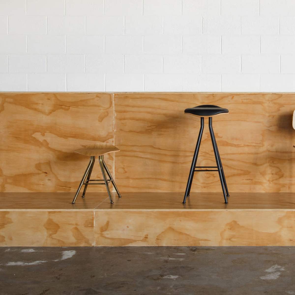 A simple wooden platform with three stools of different designs and a potted plant against a white wall.