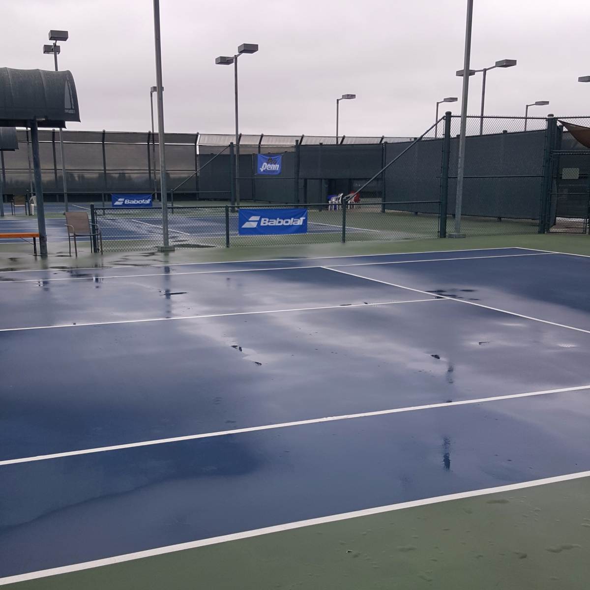 A set of wet tennis courts with blue and green surfaces, surrounded by poles with lights and a covered structure in the background.