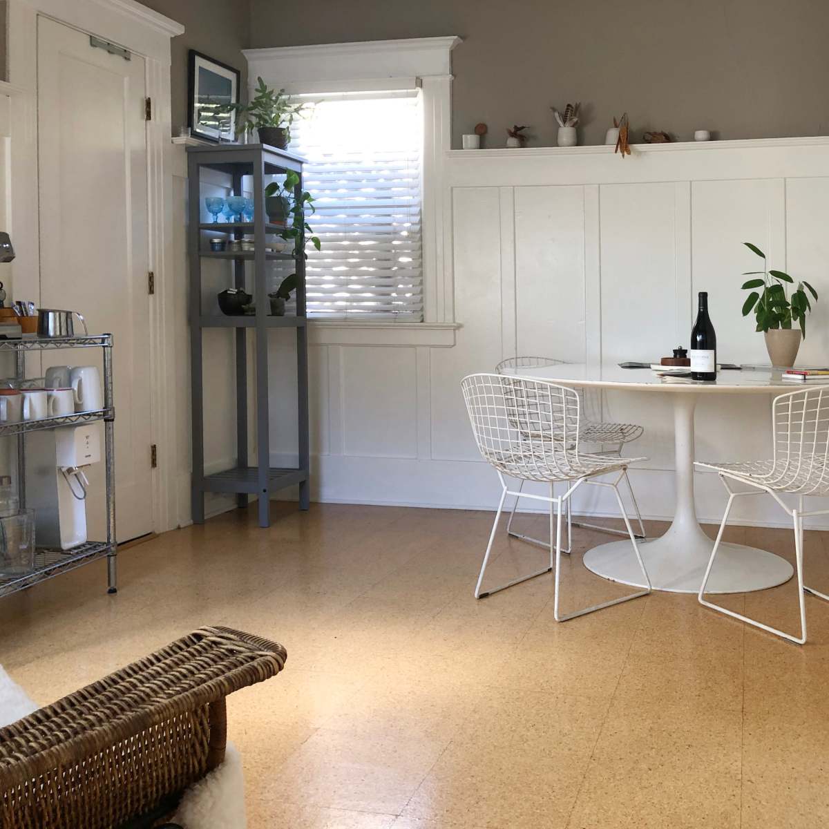 A small dining area features a round white table with two wire chairs, a bottle of wine, and a potted plant, complemented by a shelving unit and light from a window.