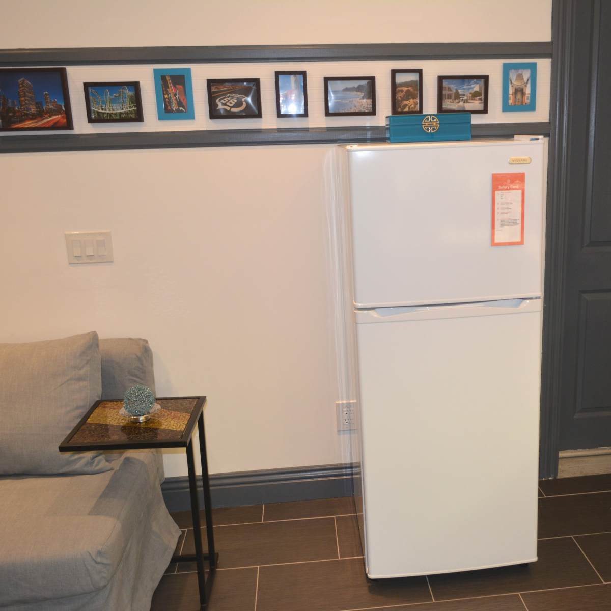 A white refrigerator positioned next to a couch and framed pictures on the wall above.