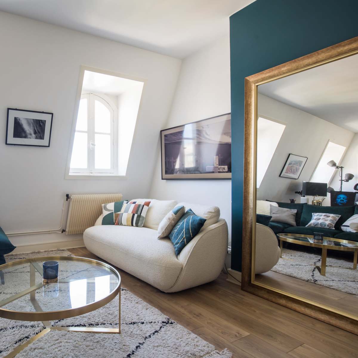 A modern living room featuring a blue sofa, a cream-colored couch, a glass coffee table, and a large mirror reflecting the space.