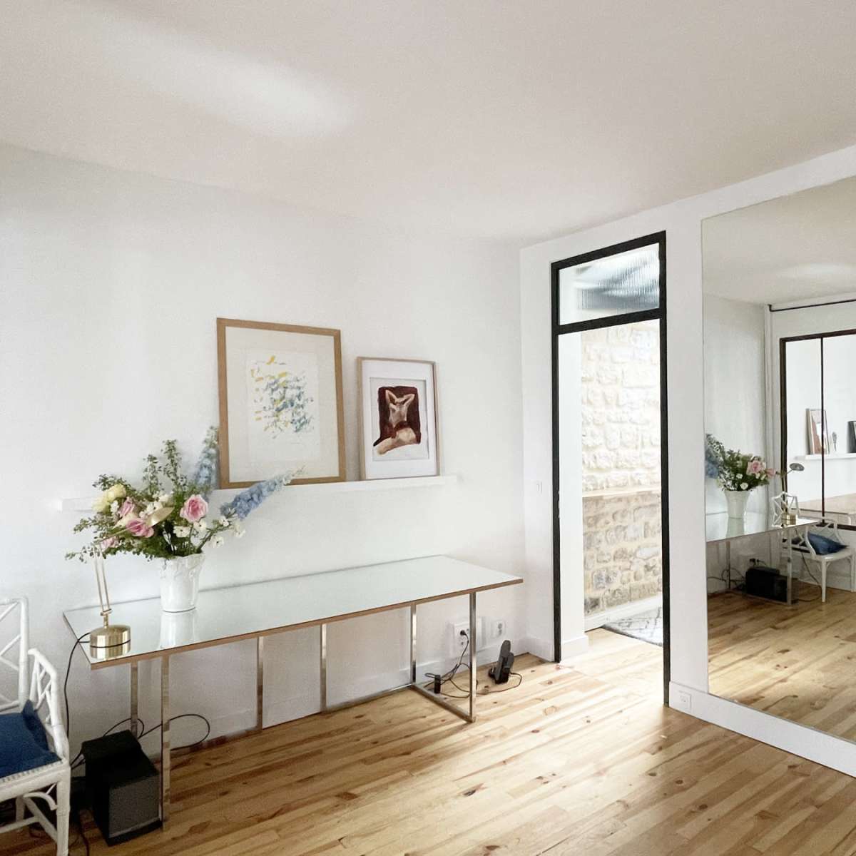 A well-lit room featuring a glass table with a floral arrangement, framed artwork on the wall, and a large mirror reflecting an adjacent space with stone walls.