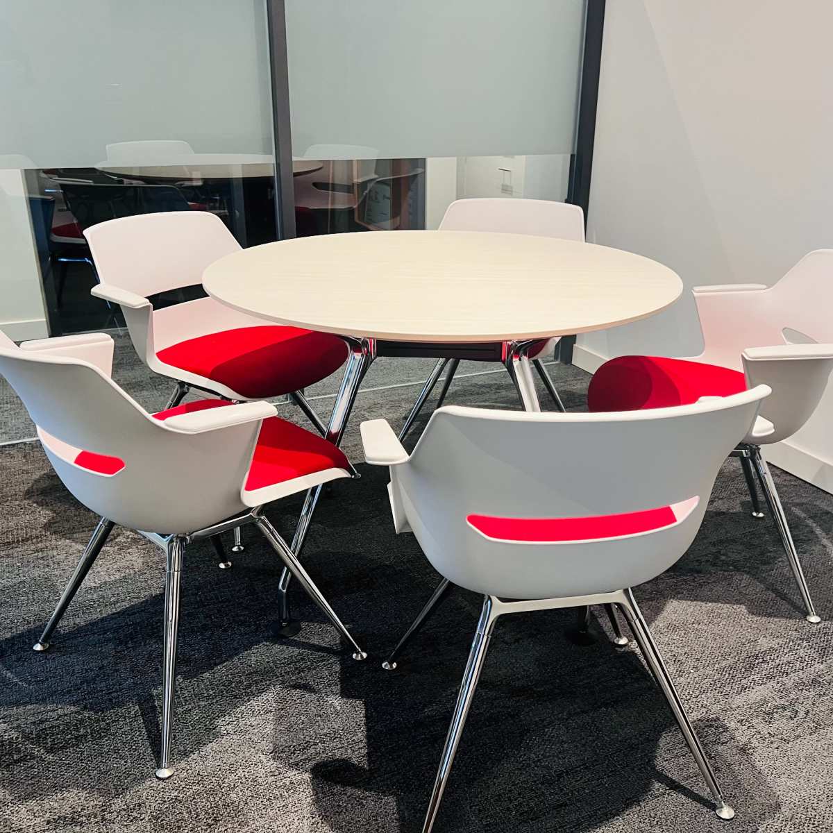 A round table surrounded by five white chairs with red seat cushions sits in a modern office setting.