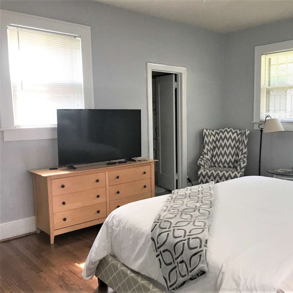 A tidy bedroom with a bed, a television on a wooden dresser, and a chair next to a lamp near the window.