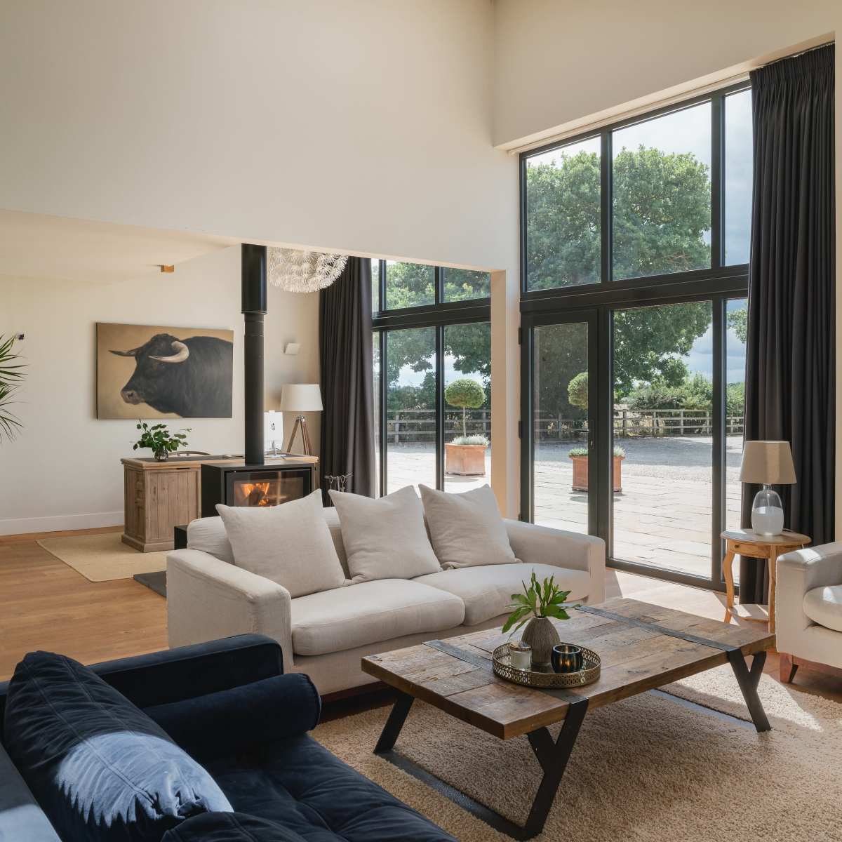 A spacious living room featuring a light-colored sofa, a dark blue couch, a wooden coffee table, and large windows with views of greenery outside.