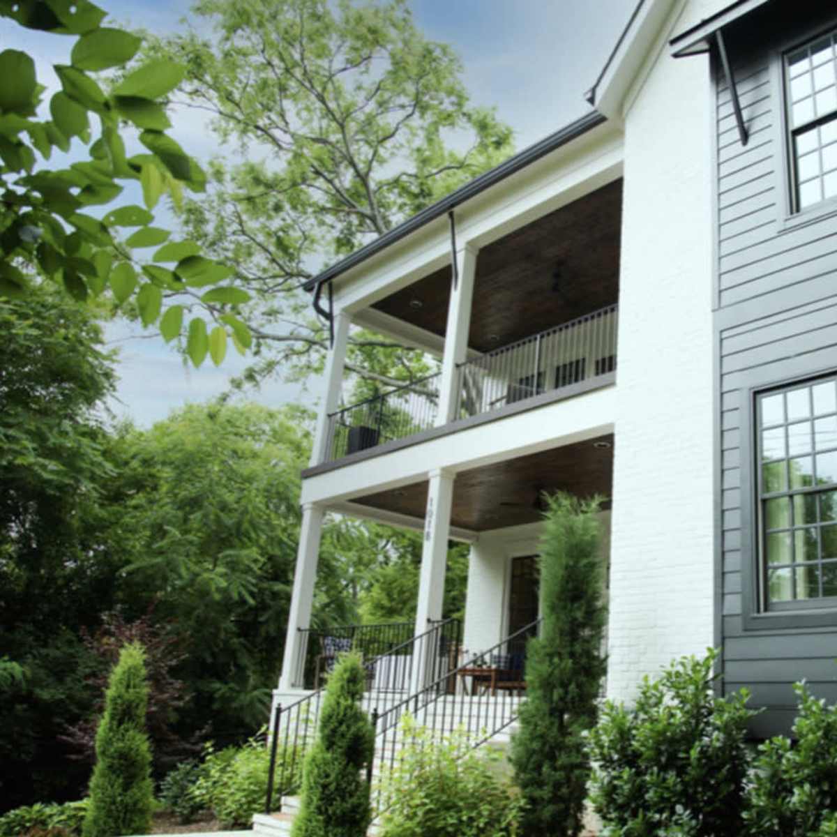A two-story house with a mix of black and white siding, featuring large windows and a covered porch surrounded by greenery.