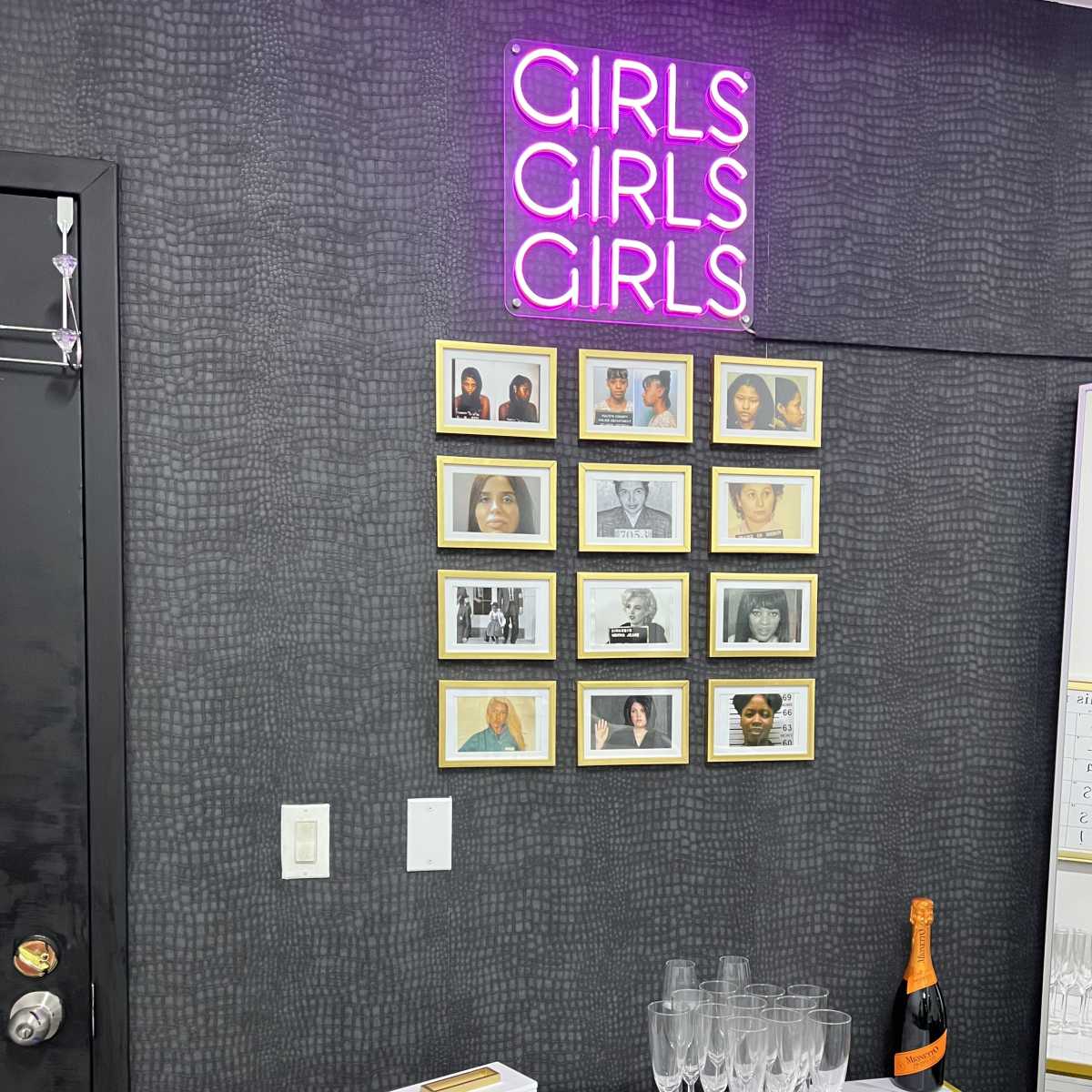 A wall displays a neon sign reading "GIRLS GIRLS GIRLS" above a collection of framed photographs, a table with champagne flutes, and a bottle of champagne.