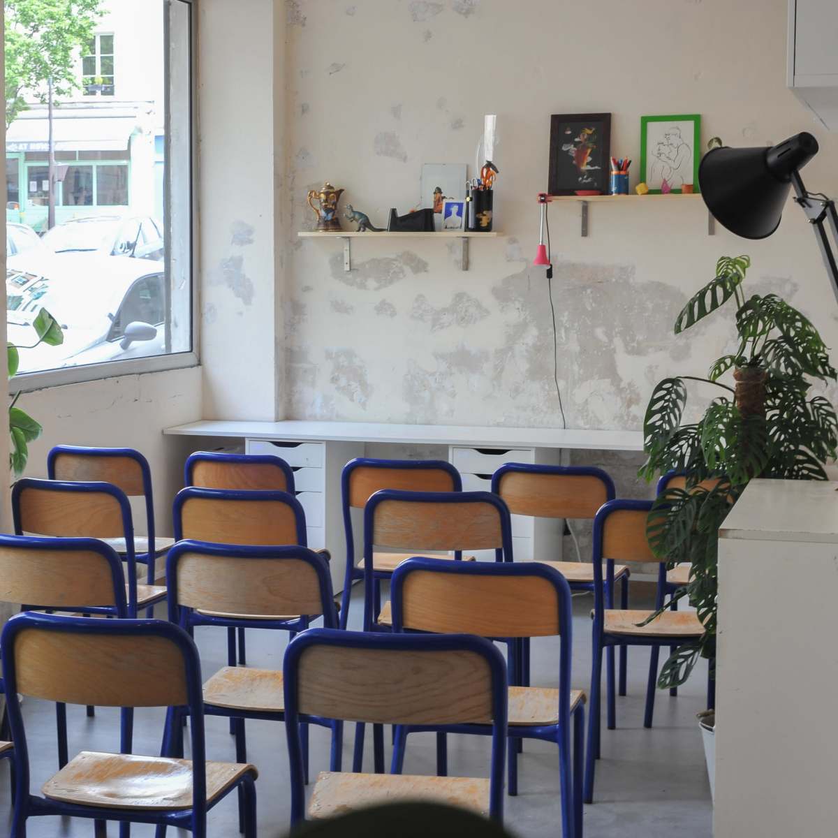 A minimalist room with wooden and blue metal chairs arranged in rows facing a window and a shelf displaying various items.