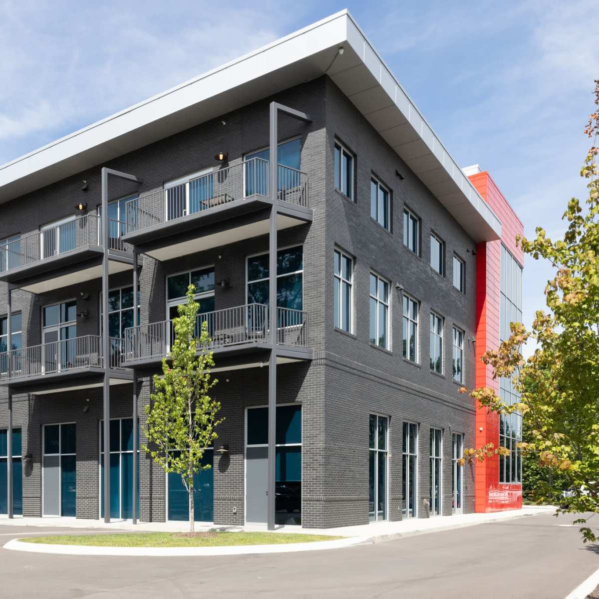 A modern three-story commercial building with a grey brick facade, balconies, large windows, and a section painted in red.