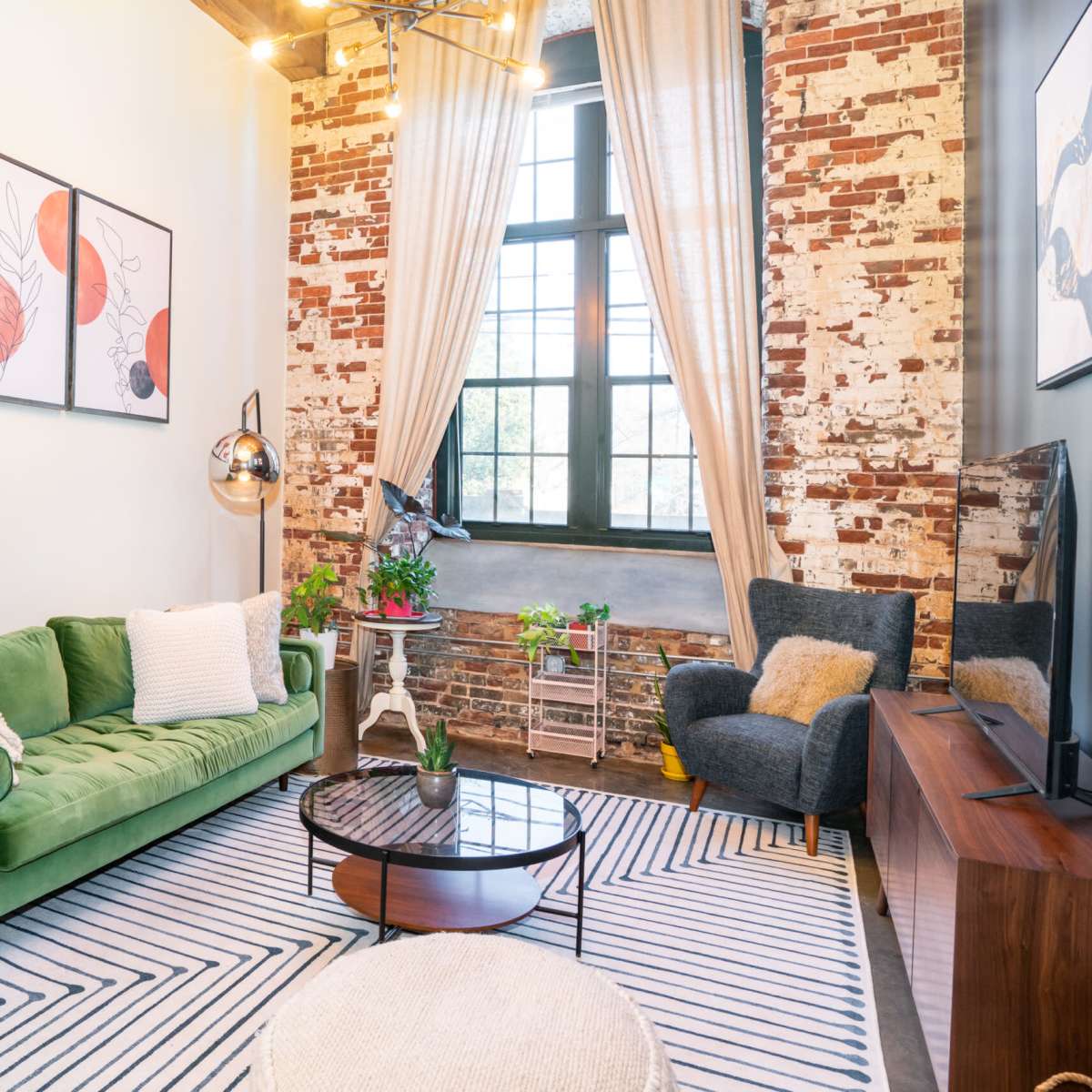 A modern living room with a green sofa, a circular wooden coffee table, and exposed brick walls, complemented by large windows and decorative artwork.