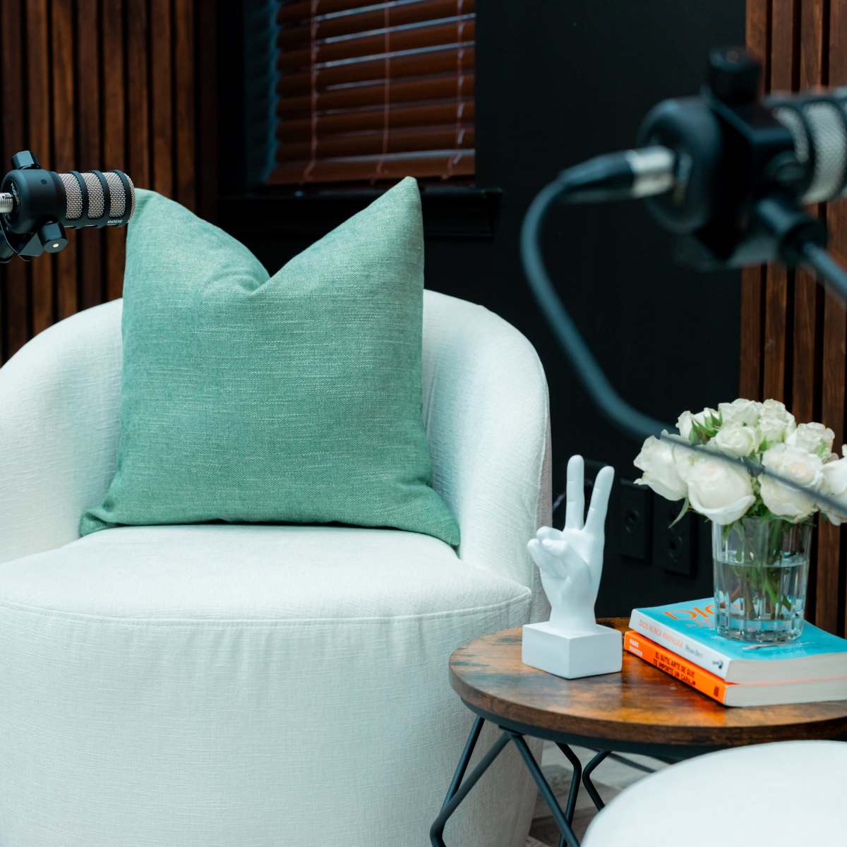 A cozy seating area features two white armchairs with a green pillow, a small table holding a vase of flowers, and two microphones positioned nearby.