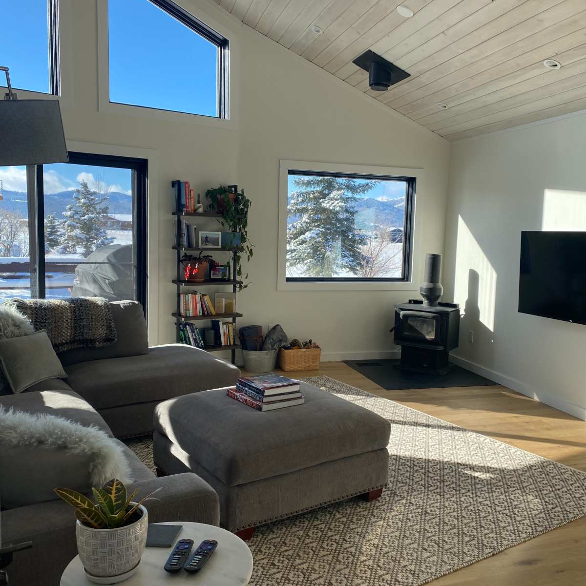 The living room features a gray sectional sofa, a wall-mounted television, a wood stove, and large windows displaying a view of mountains and trees.
