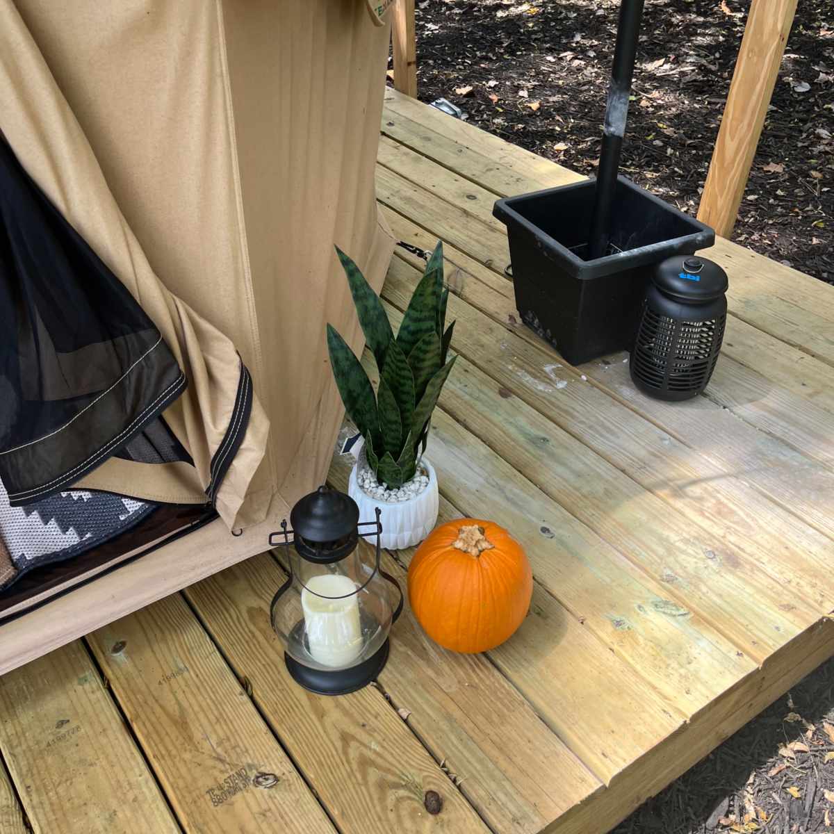 A tent is set up on a wooden deck, accompanied by a lantern with a candle, a potted plant, and a small pumpkin.