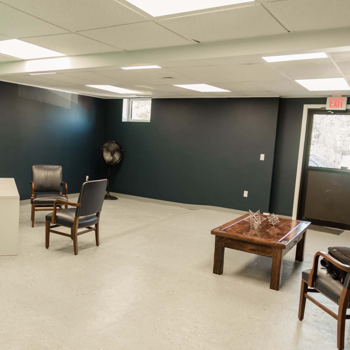 A sparsely furnished room with a white table, several chairs, and a large window.