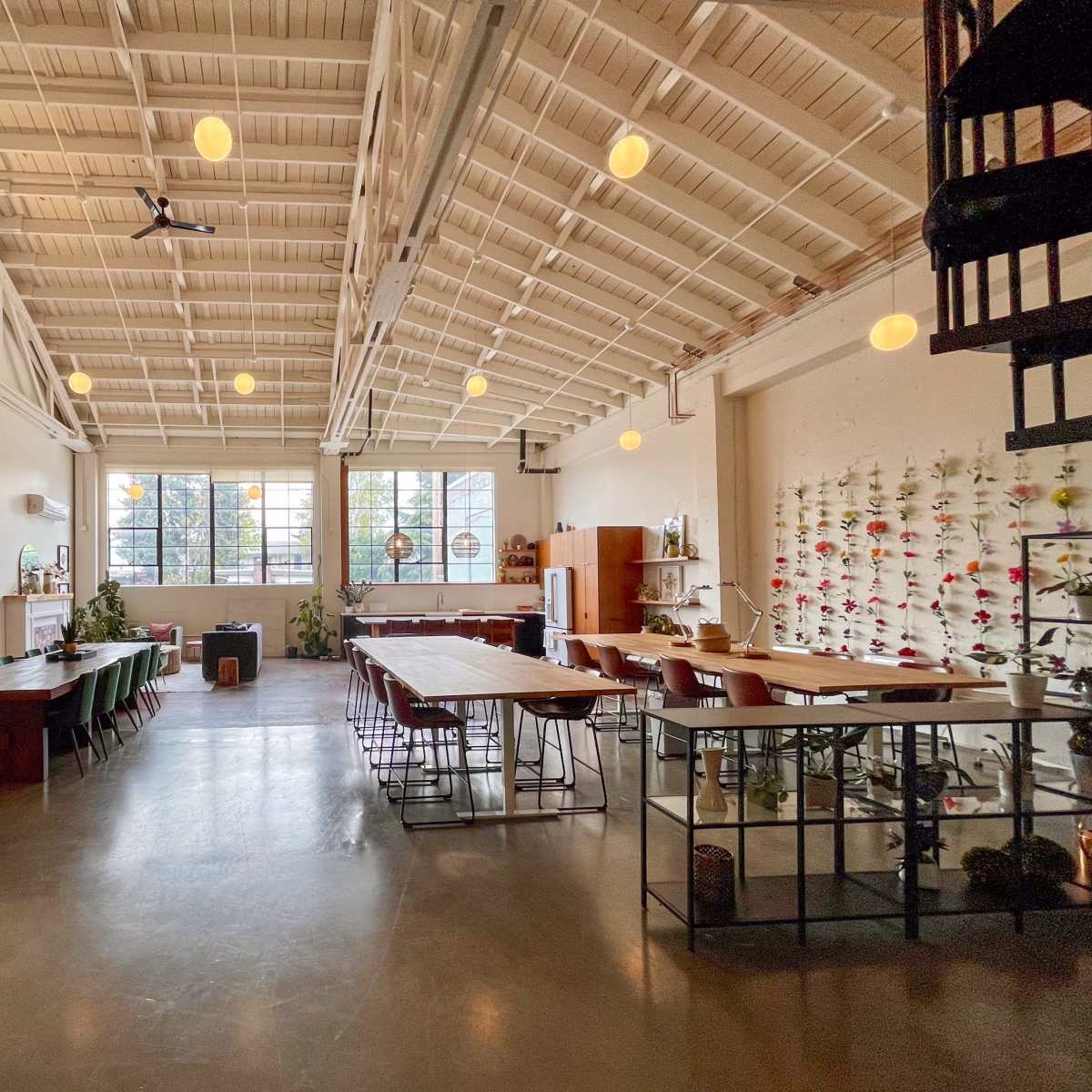 A spacious room with high ceilings, featuring large windows, a mix of dining and work tables, and a vertical garden on one wall.