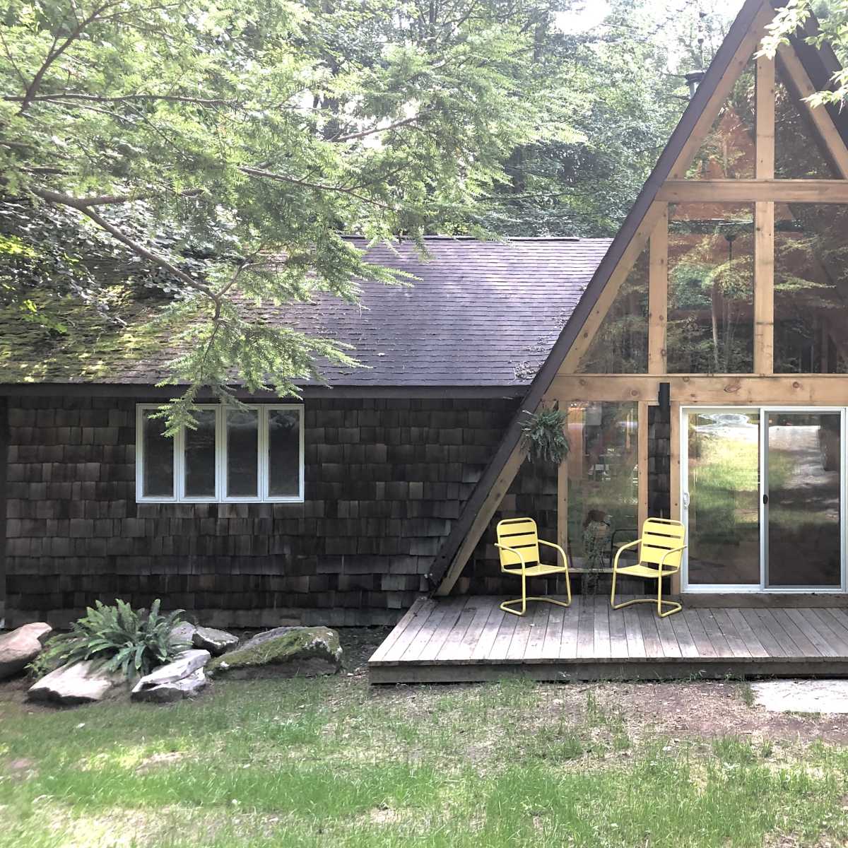 A wooden A-frame cabin with large front windows features a small deck and two yellow chairs.