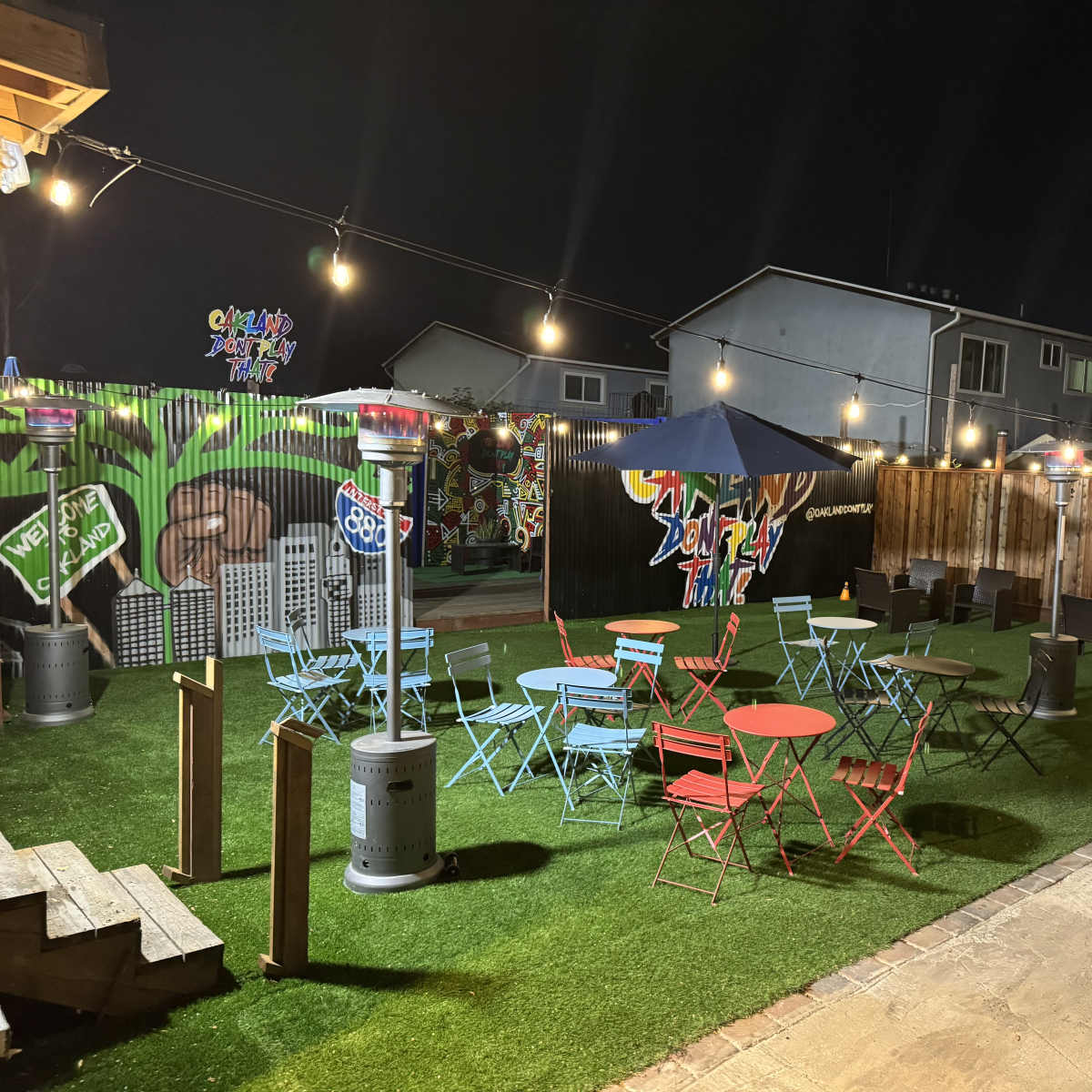 A backyard area at night with colorful tables and chairs, patio heaters, and string lights illuminating a vibrant mural on the wall.
