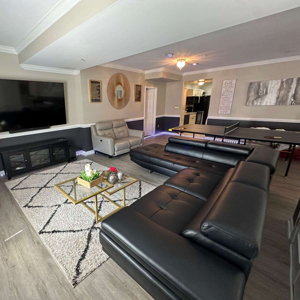 The scene features a modern living room with a large dark sectional sofa, a gold coffee table, a mounted flat-screen TV, and minimalistic decor against light-colored walls.