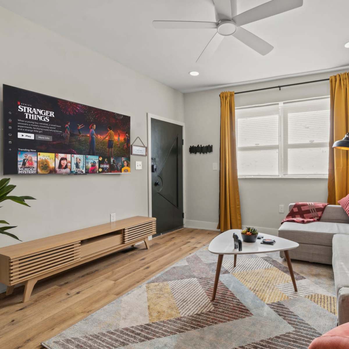 A modern living room features a sectional couch, a coffee table with remote controls, a large TV mounted on the wall displaying a streaming service, and bright yellow curtains framing a window.