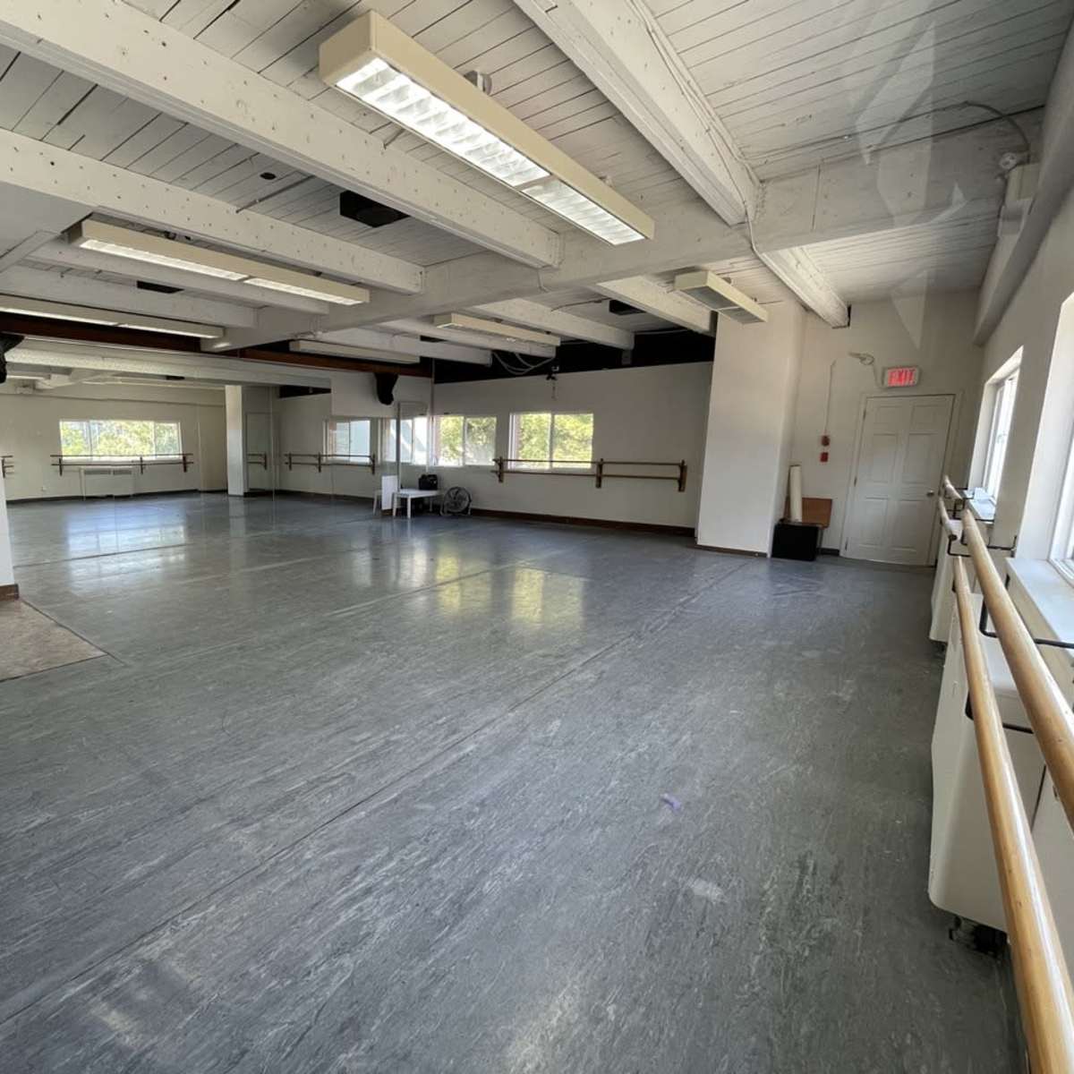 A spacious, well-lit dance studio with grey flooring, large windows, and mirrored walls.