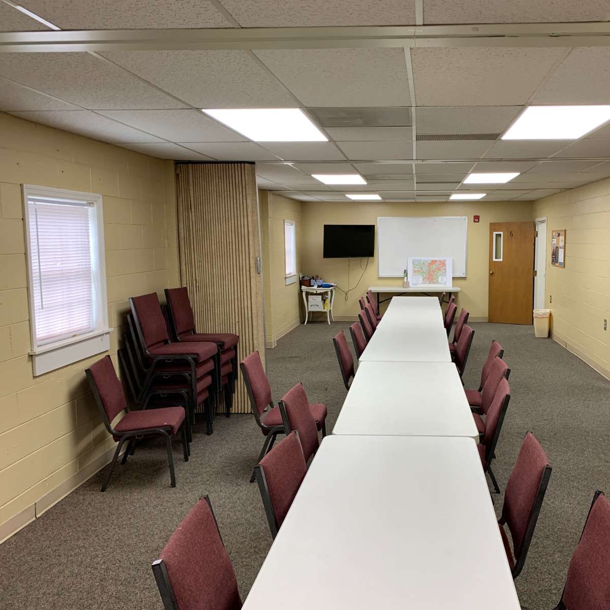 A long, rectangular table with chairs on either side is set up in a spacious room featuring beige walls, multiple light fixtures, and two doors.
