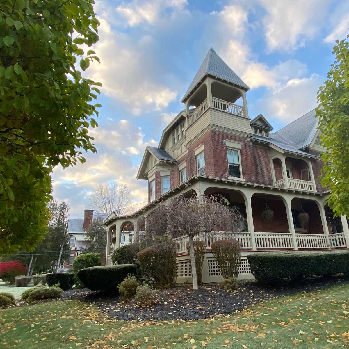 A large, historic brick house with a turret and a wraparound porch is surrounded by well-manicured lawns and trees.
