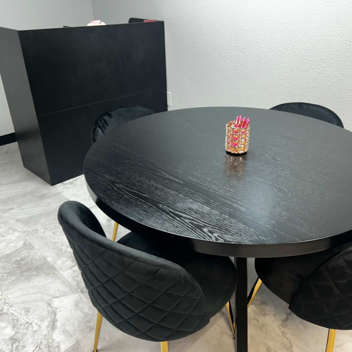 A round black dining table with four black chairs and a decorative piece in the center is positioned in a room with light-colored flooring and a black reception desk in the background.