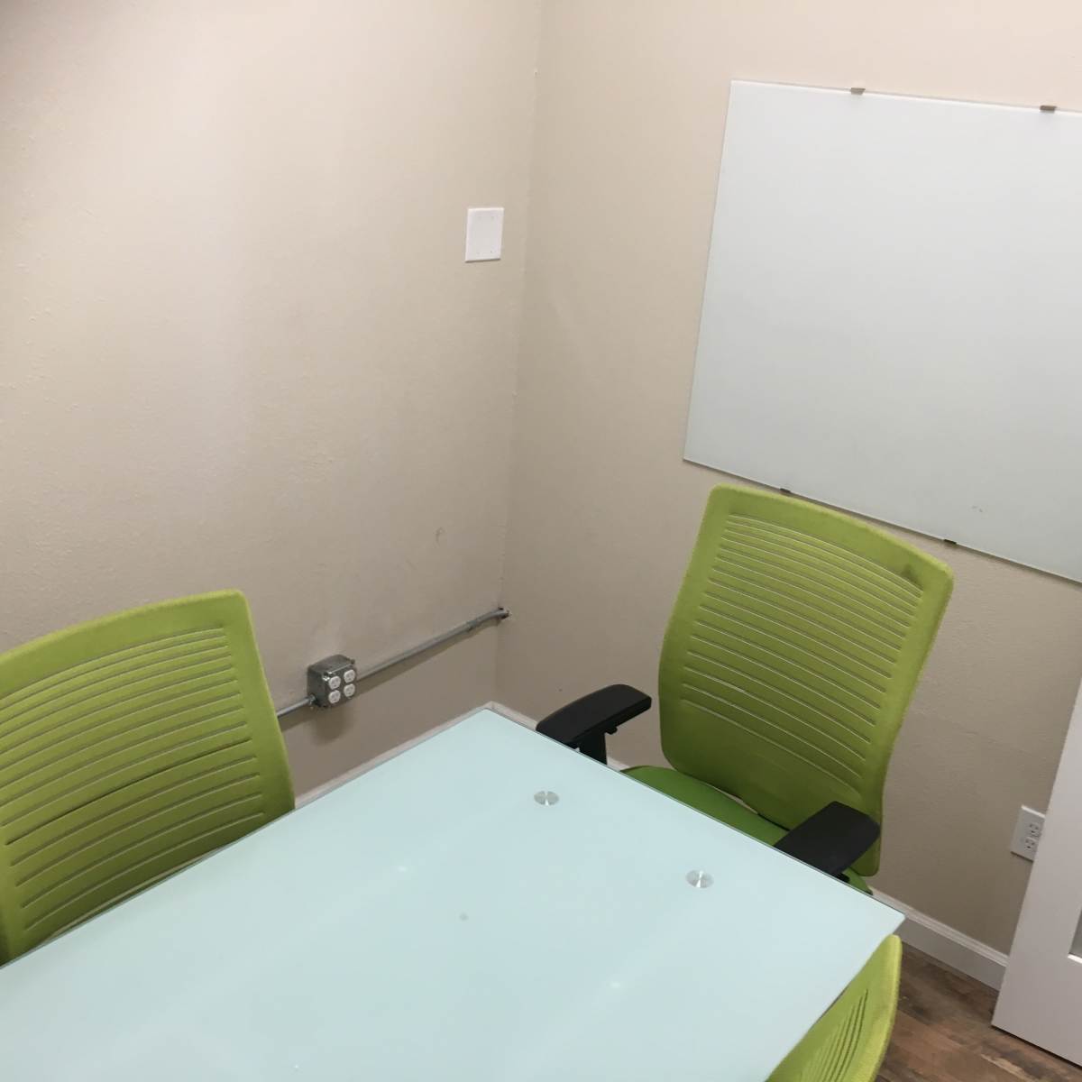 A small meeting room with a glass table, two green chairs, and a whiteboard attached to the wall.