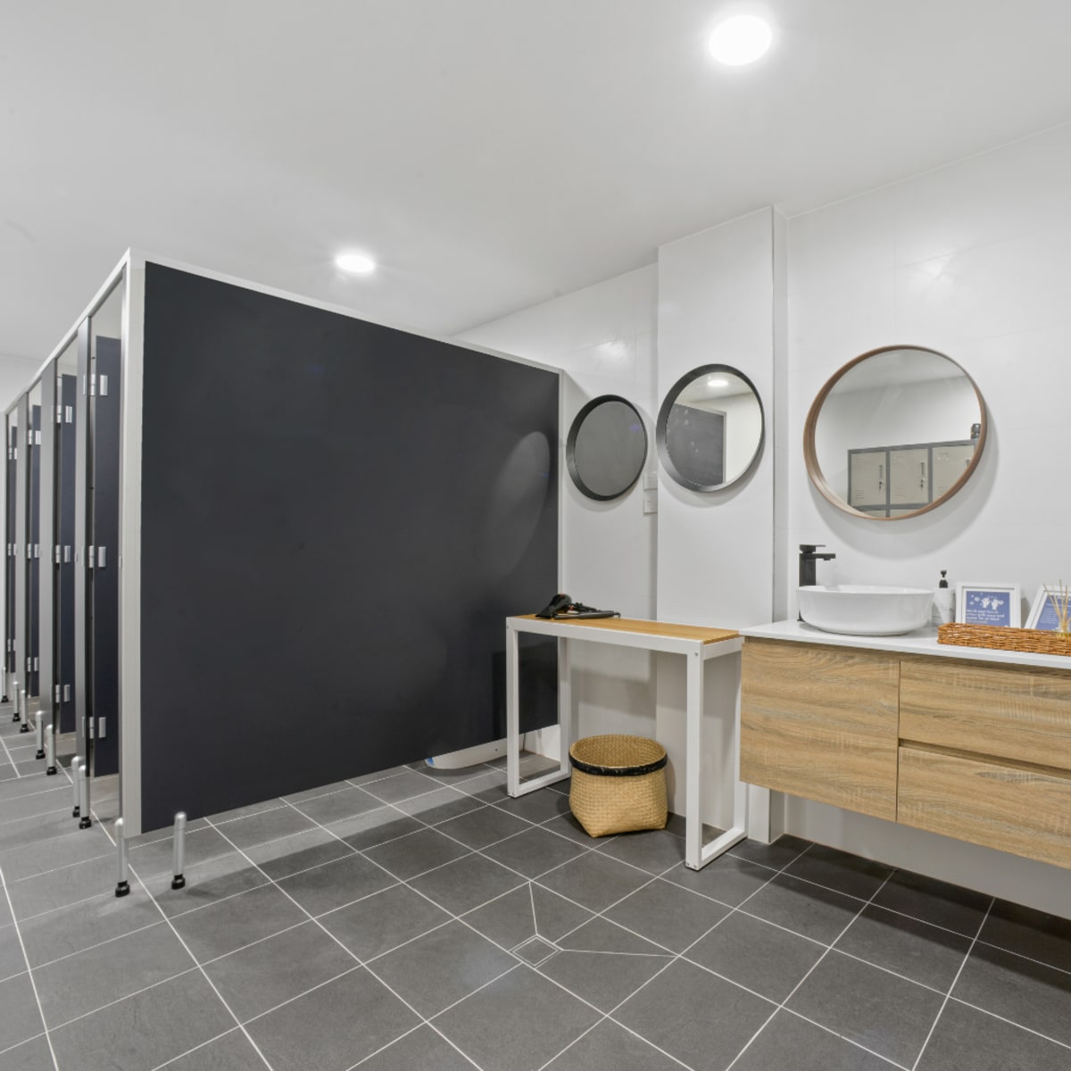 A modern bathroom with private stalls, round mirrors, and a wooden vanity with two sinks.
