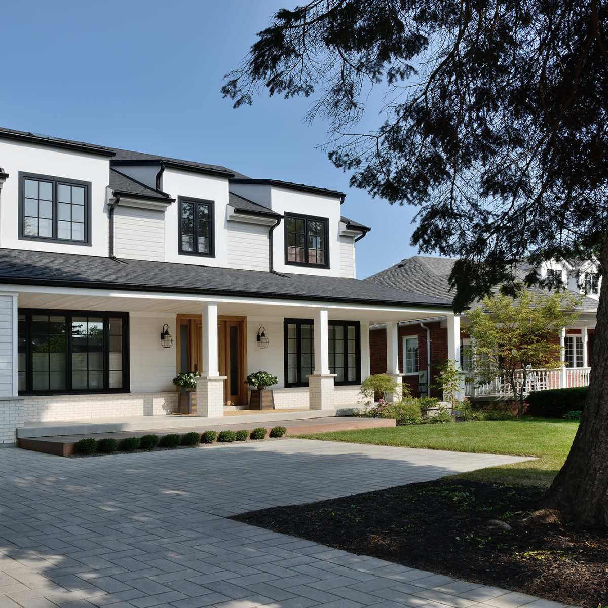 A modern two-story house with white siding, black windows, and a dark roof is set on a neatly paved driveway surrounded by a well-maintained lawn.