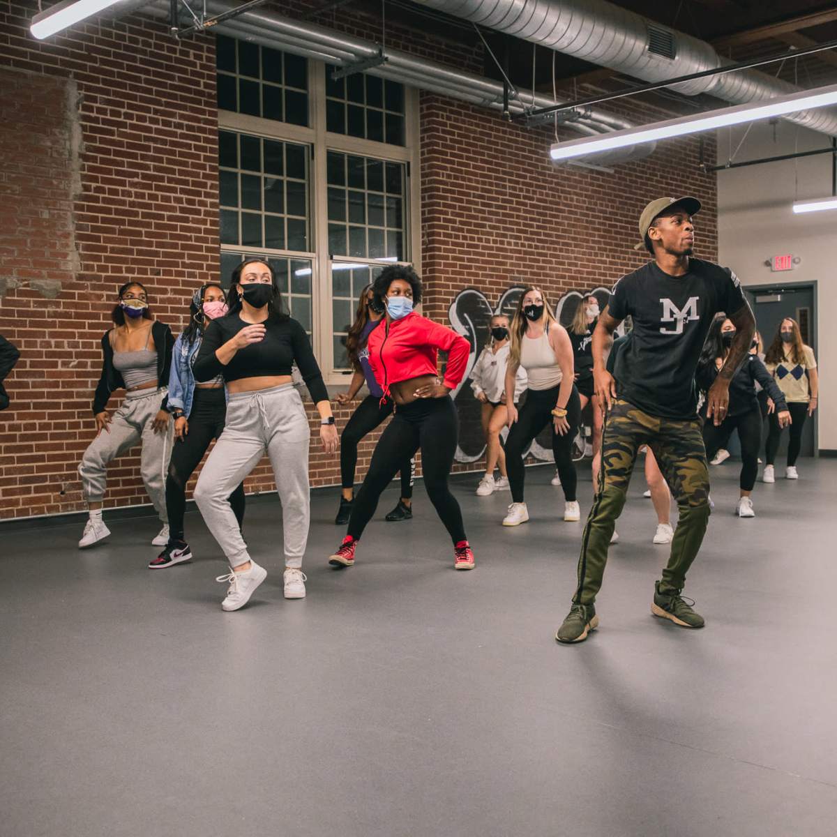 A group of dancers in workout attire perform choreographed moves in a spacious studio with brick walls and large windows.