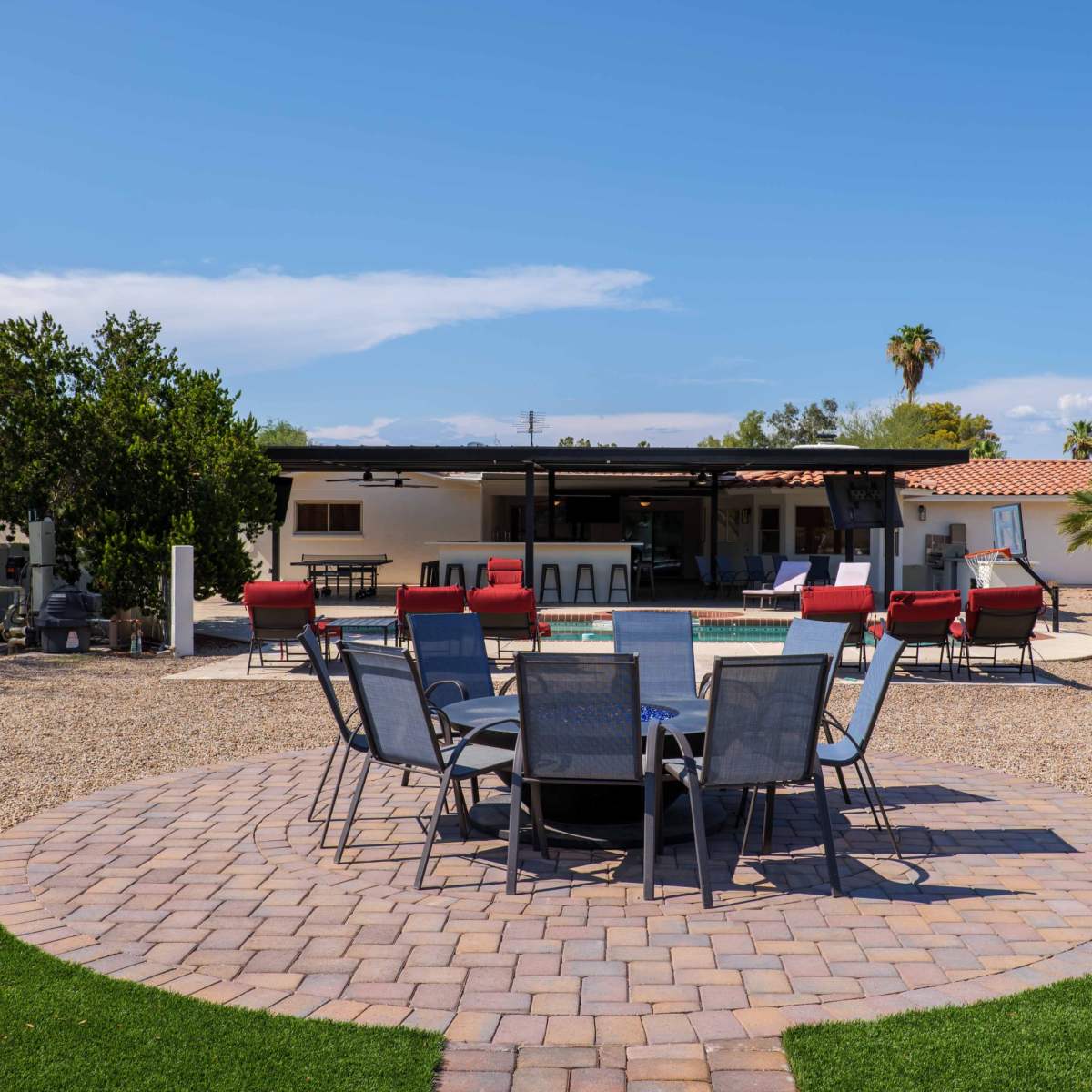 A landscaped backyard with a circular stone patio surrounded by chairs, a pool, and a house in the background.