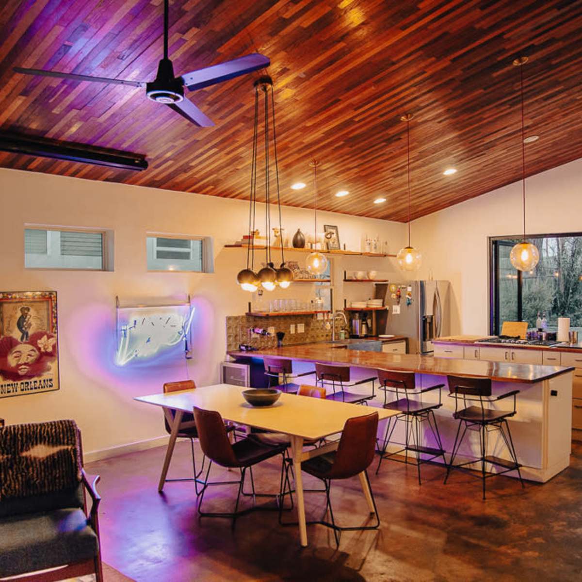 A modern kitchen and dining area features wooden beams, a large island, and a blend of pendant lighting, with a view of an outdoor space through large glass doors.