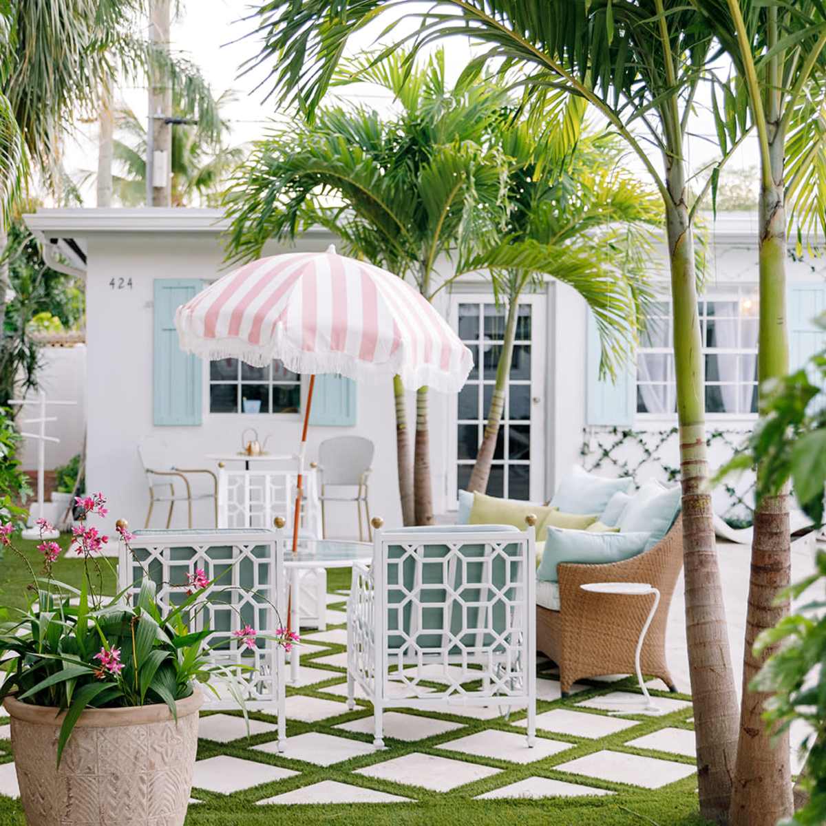 A garden area with white furniture, a pink and white striped umbrella, and various tropical plants surrounding a small table and chairs.