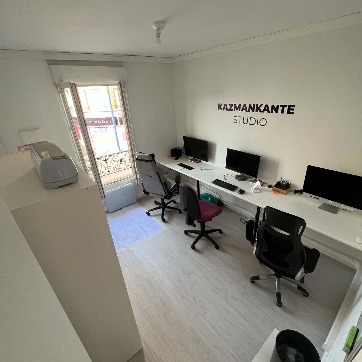 A minimalist office space with several desks, computers, and a window overlooking a balcony, featuring the name "KAZMANKANTE STUDIO" on the wall.
