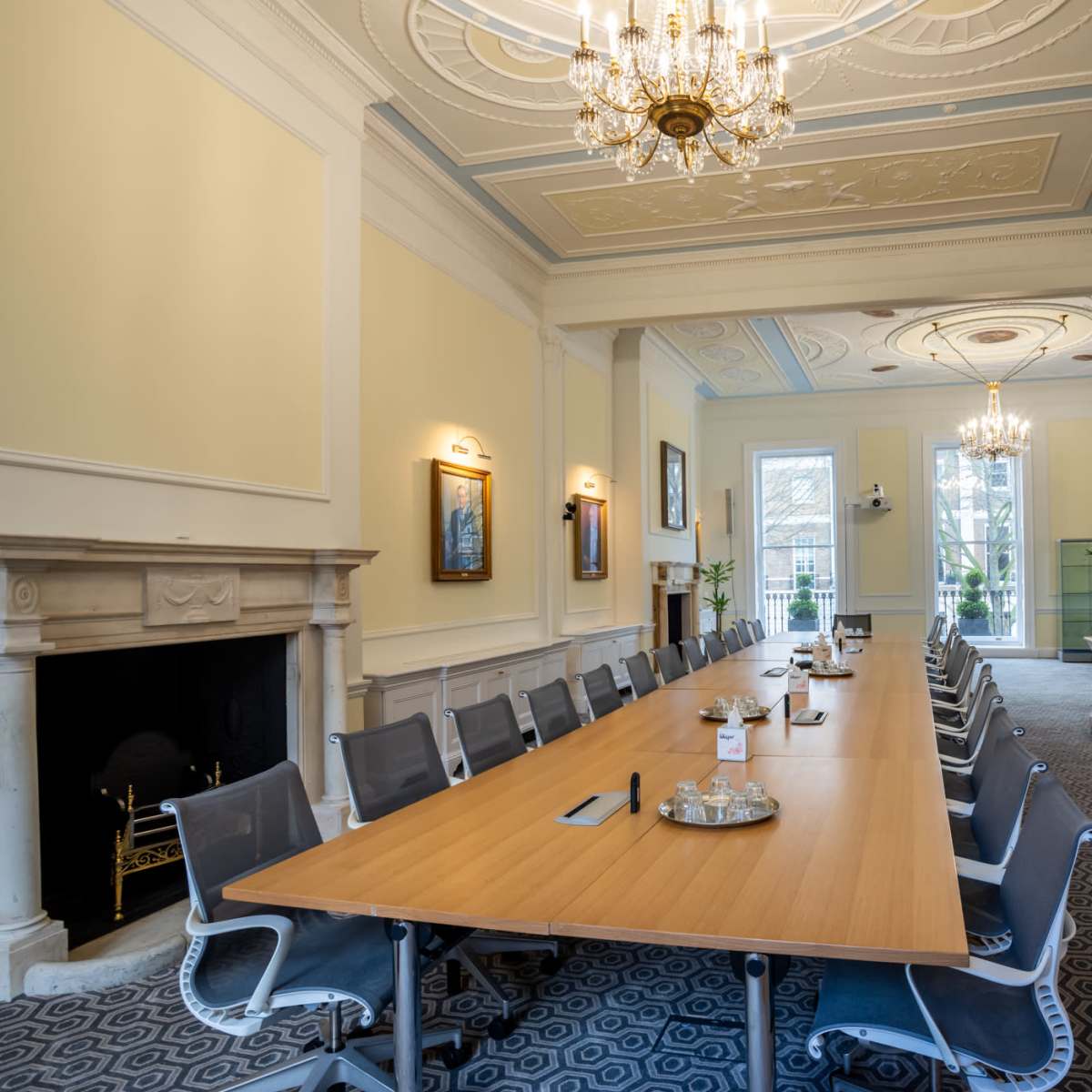 A spacious conference room features a long wooden table surrounded by modern chairs, with large windows allowing natural light and ornate chandeliers overhead.