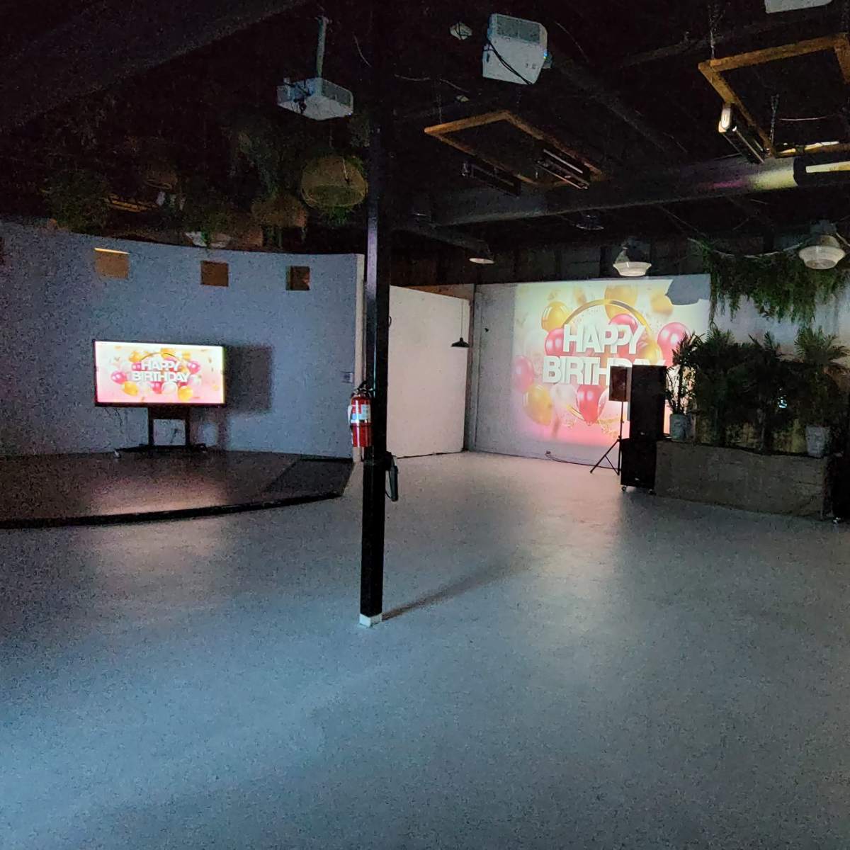 A spacious event room with a large "Happy Birthday" projection on the wall, a television, and sound equipment arranged near a planter.