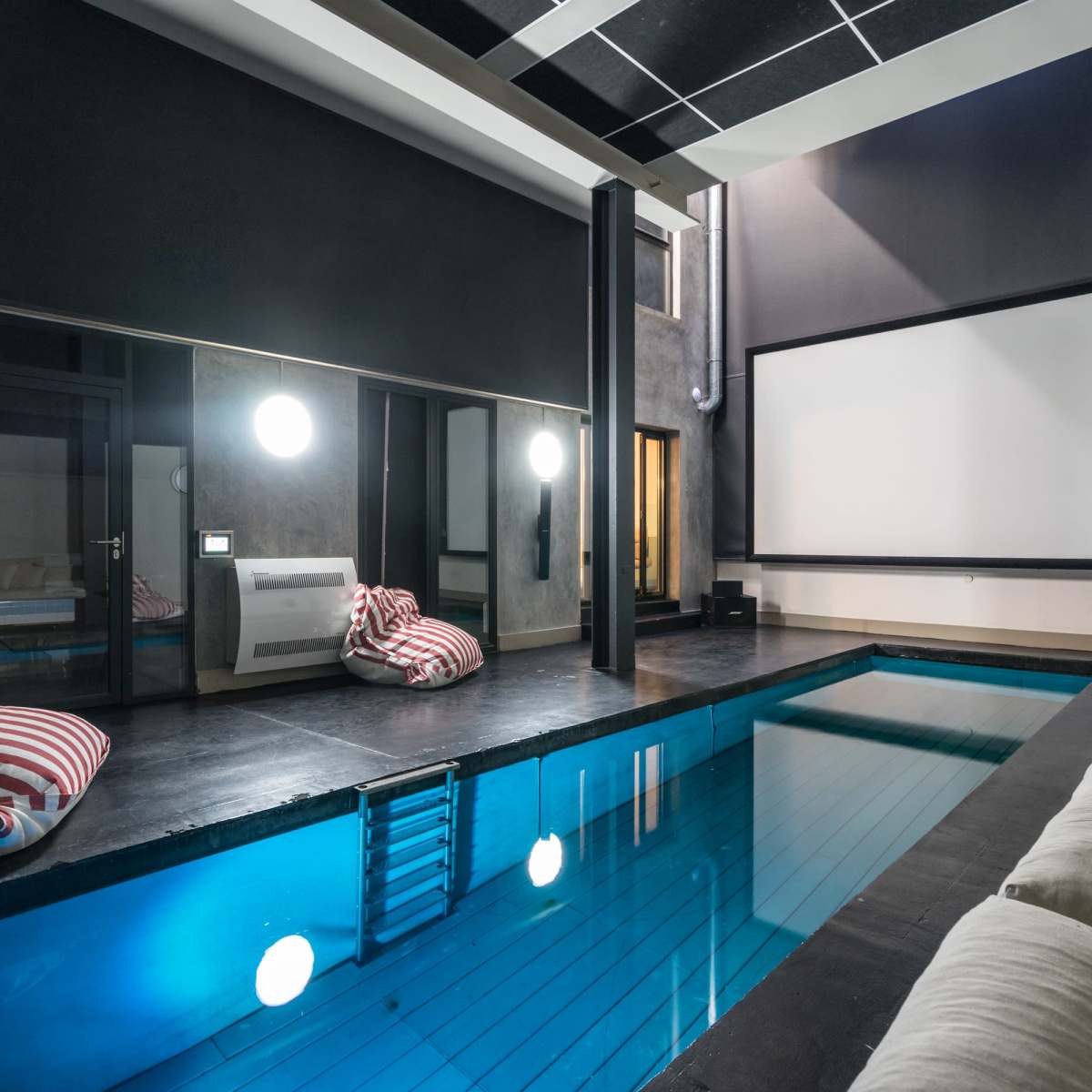 An indoor pool area with a large movie screen, bean bag chairs, and a mirrored wall reflecting the pool.